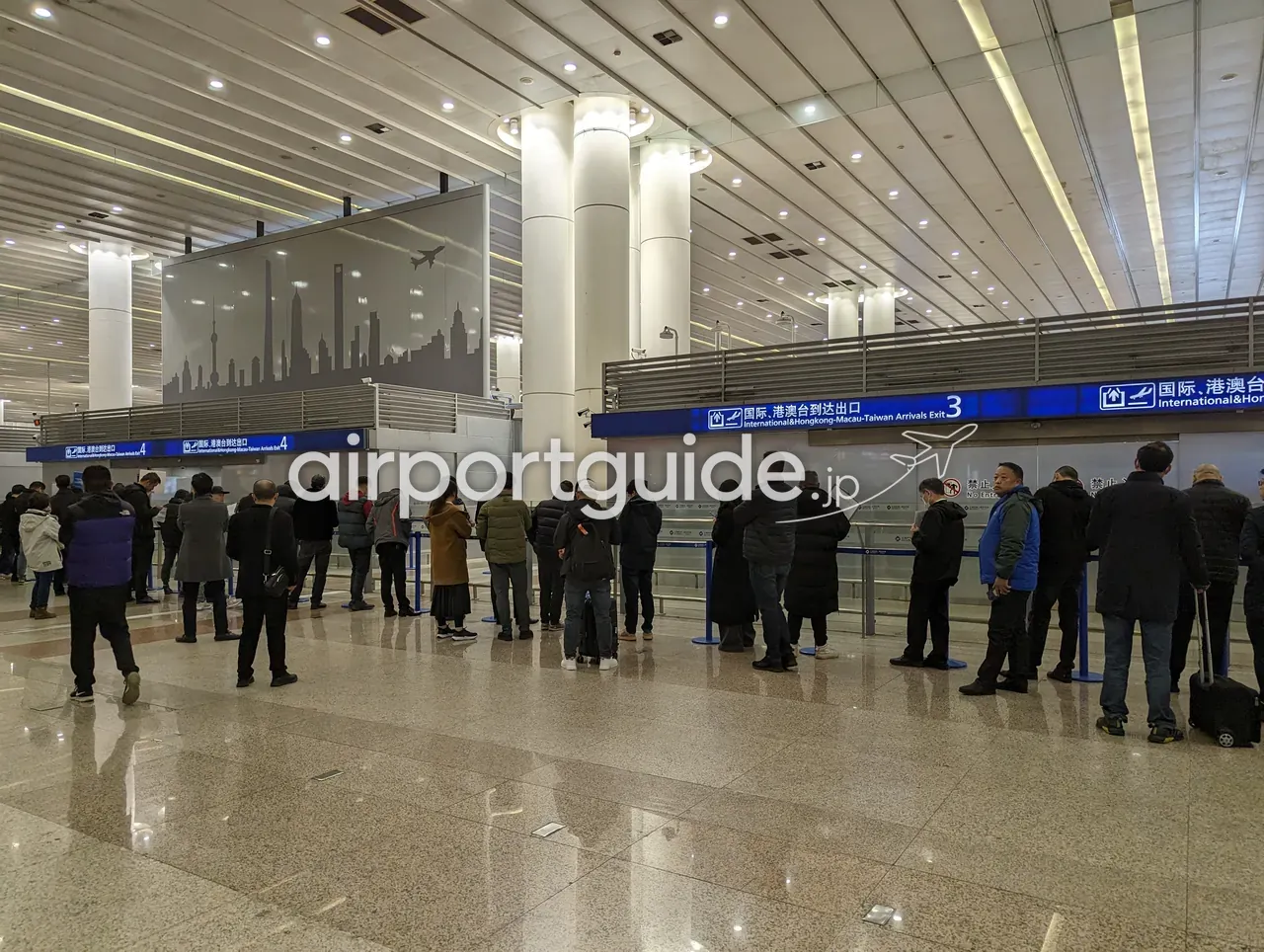 上海浦東空港の国際到着口。『国际、港澳台到达出口 International&Hongkong-Macau-Taiwan Arrivals Exit 3／4』の青い看板と、上海の摩天楼（東方明珠塔などのシルエット）を描いた壁面、出迎えの人だかり。