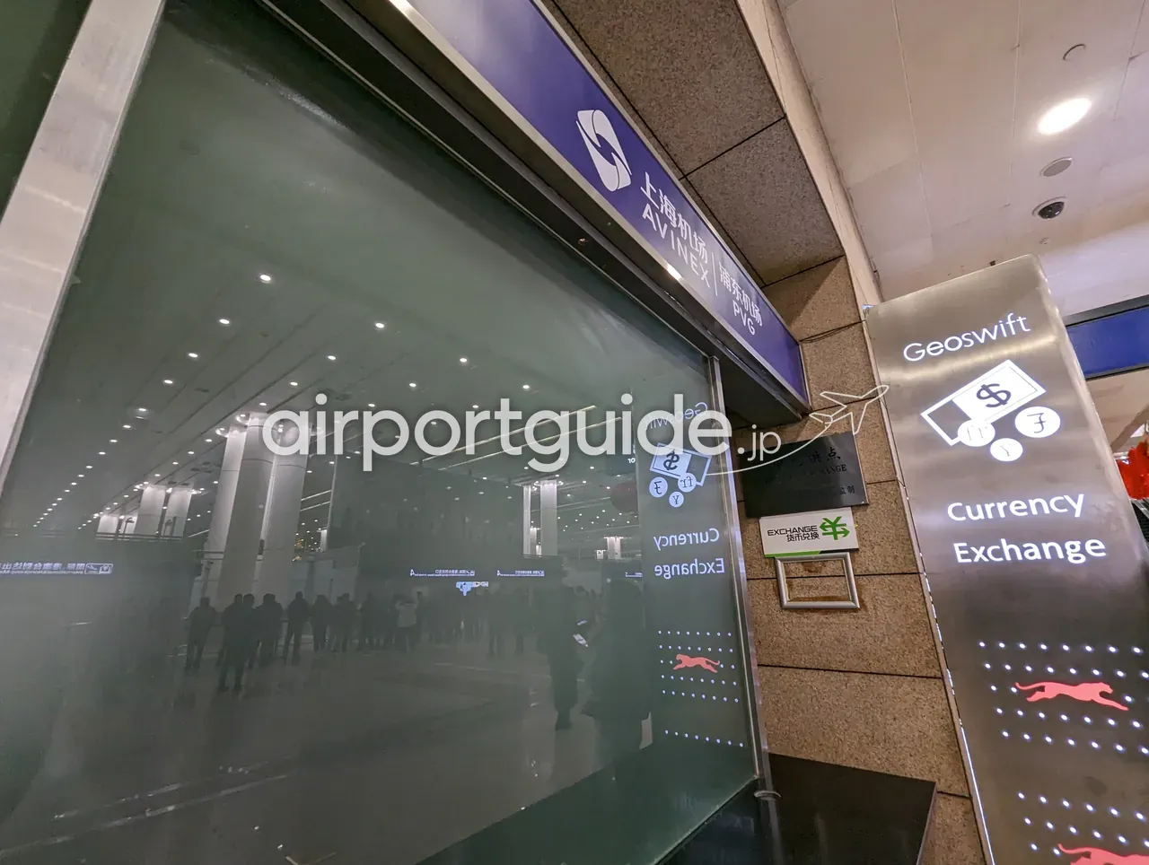 上海浦東空港（PVG）の到着階。ガラスの上に「上海机场 AVINEX｜浦东机场 PVG」のサイン、右にステンレスの案内塔「Geoswift Currency Exchange 货币兑换」（各国通貨のアイコンと走る犬のマーク）。ガラス越しに到着ホールと並ぶ人々が映っている。