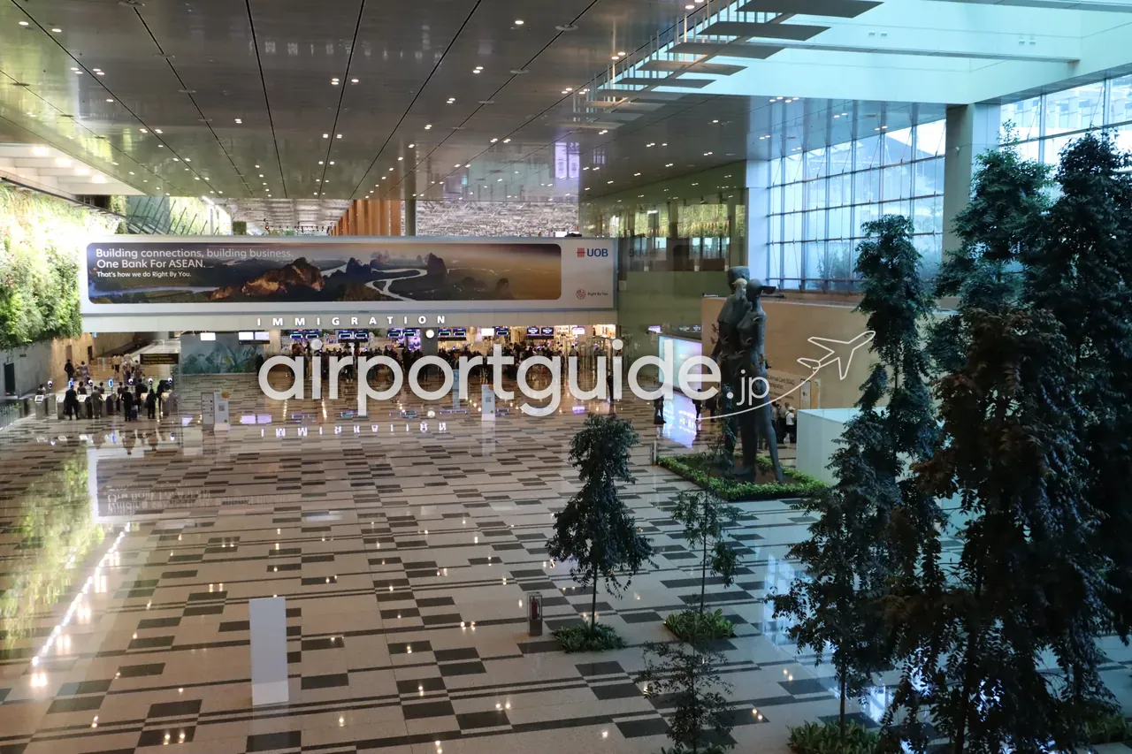 チャンギ空港の到着イミグレーションホールを高所から俯瞰。「IMMIGRATION」のサインの下に入国審査カウンターが並び、両脇は緑化壁と樹木。