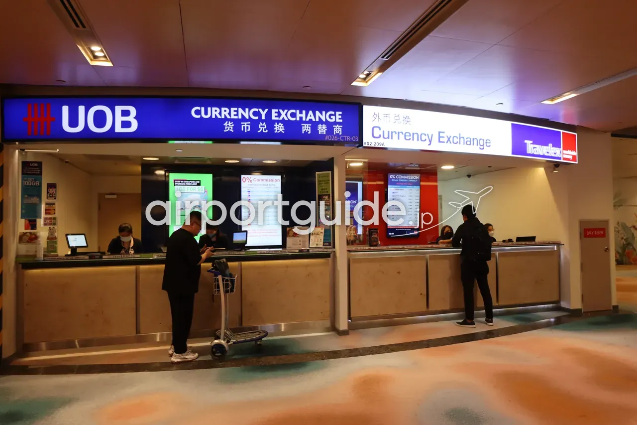 チャンギ空港の両替カウンター。左が青い「UOB CURRENCY EXCHANGE 货币兑换 两替商」、右が「Currency Exchange」とTravelexのロゴ。どちらの窓口にも「0% Commission」のレートボードがあり、スーツケースを持つ旅行者が両替の手続きをしている。