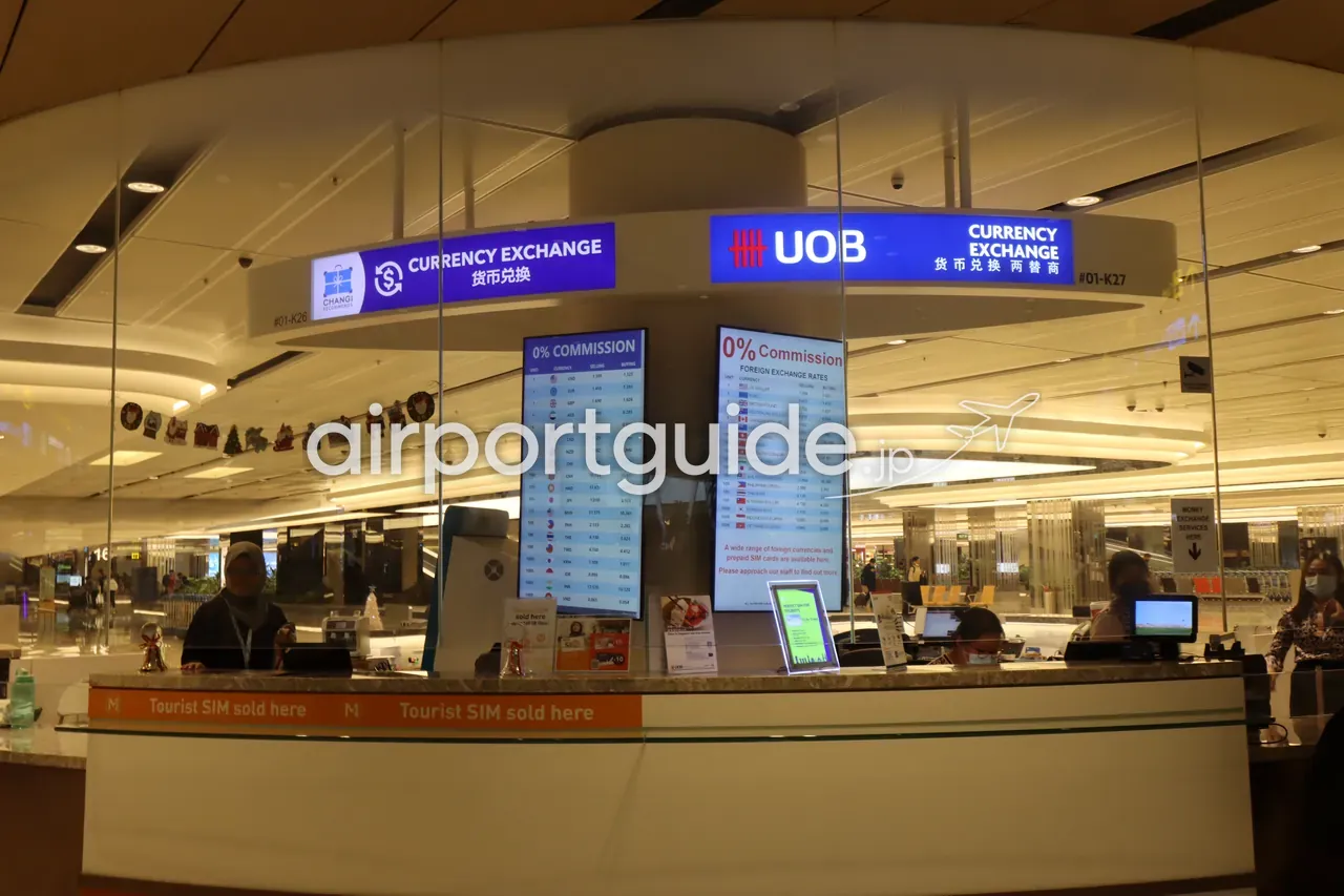 チャンギ空港の円形の両替カウンター。左に『CHANGI RECOMMENDS Currency Exchange 货币兑换 #01-K26』、右に『UOB CURRENCY EXCHANGE 货币兑换 两替商 #01-K27』の青い看板。中央に2枚の『0% COMMISSION』のデジタルレートボードが並び、カウンター前面に『Tourist SIM sold here』の帯、係員が業務中。