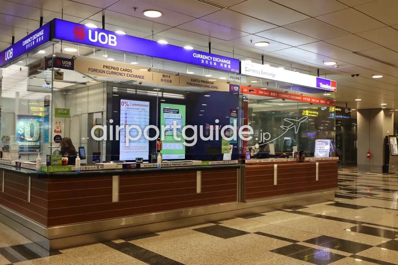 チャンギ空港のUOB両替カウンター。青い看板に『UOB CURRENCY EXCHANGE』、下段に『PREPAID CARDS／FOREIGN CURRENCY EXCHANGE』の英・中・マレー／インドネシア語表記。窓口に『0% Commission』のレートボードとSIM広告、右奥に『Ride-Hailing』『Bus to City』『Shops & 24hr Food Court』の黄色い方向サイン。隣にTravelexのカウンター。