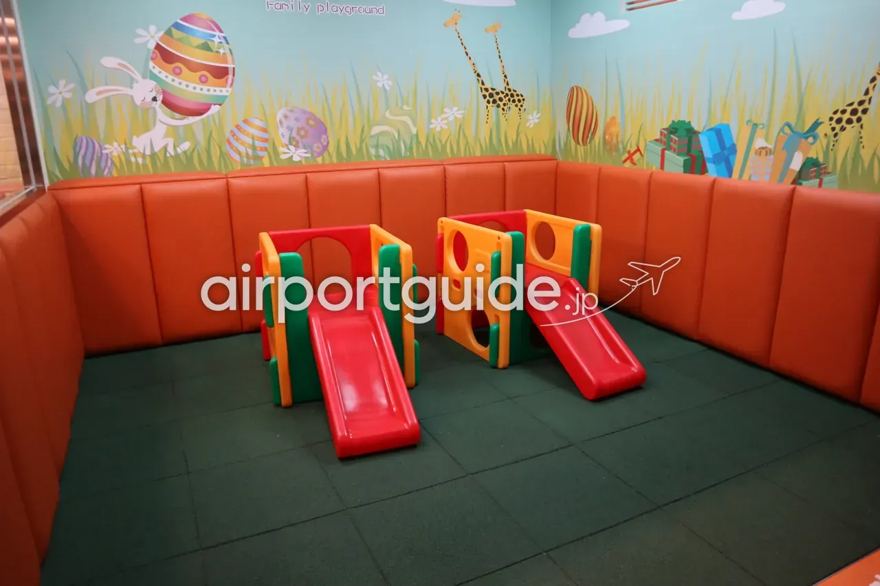 桃園空港のキッズプレイエリア「Family playground」。オレンジのクッション壁と草原・キリンの壁画、赤い滑り台付きの遊具、緑のマット床。