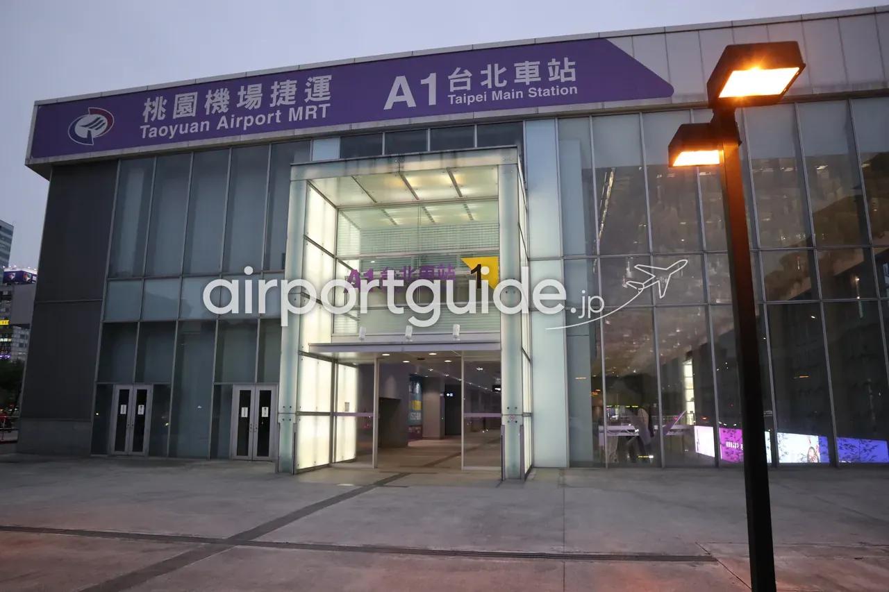 「桃園機場捷運 Taoyuan Airport MRT／A1 台北車站 Taipei Main Station」の駅入口外観。紫のサインを掲げたガラス張りの建物で、夕暮れの街灯が灯る。