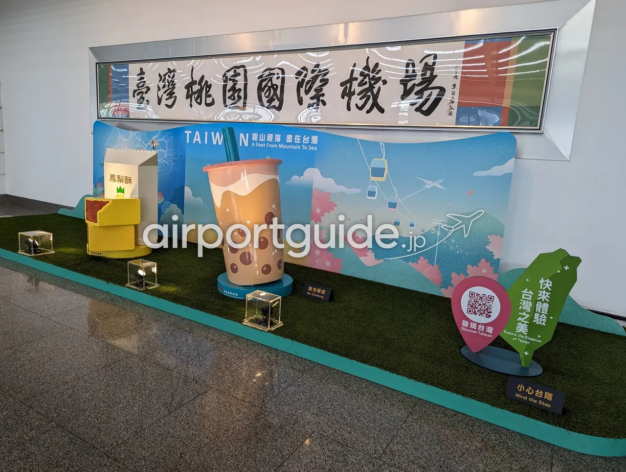 桃園空港の観光PR展示。「臺灣桃園國際機場」の書のパネルの下に、巨大なタピオカミルクティーやパイナップルケーキ（鳳梨酥）の模型と「TAIWAN A Foot From Mountain To Sea」のイラスト壁。