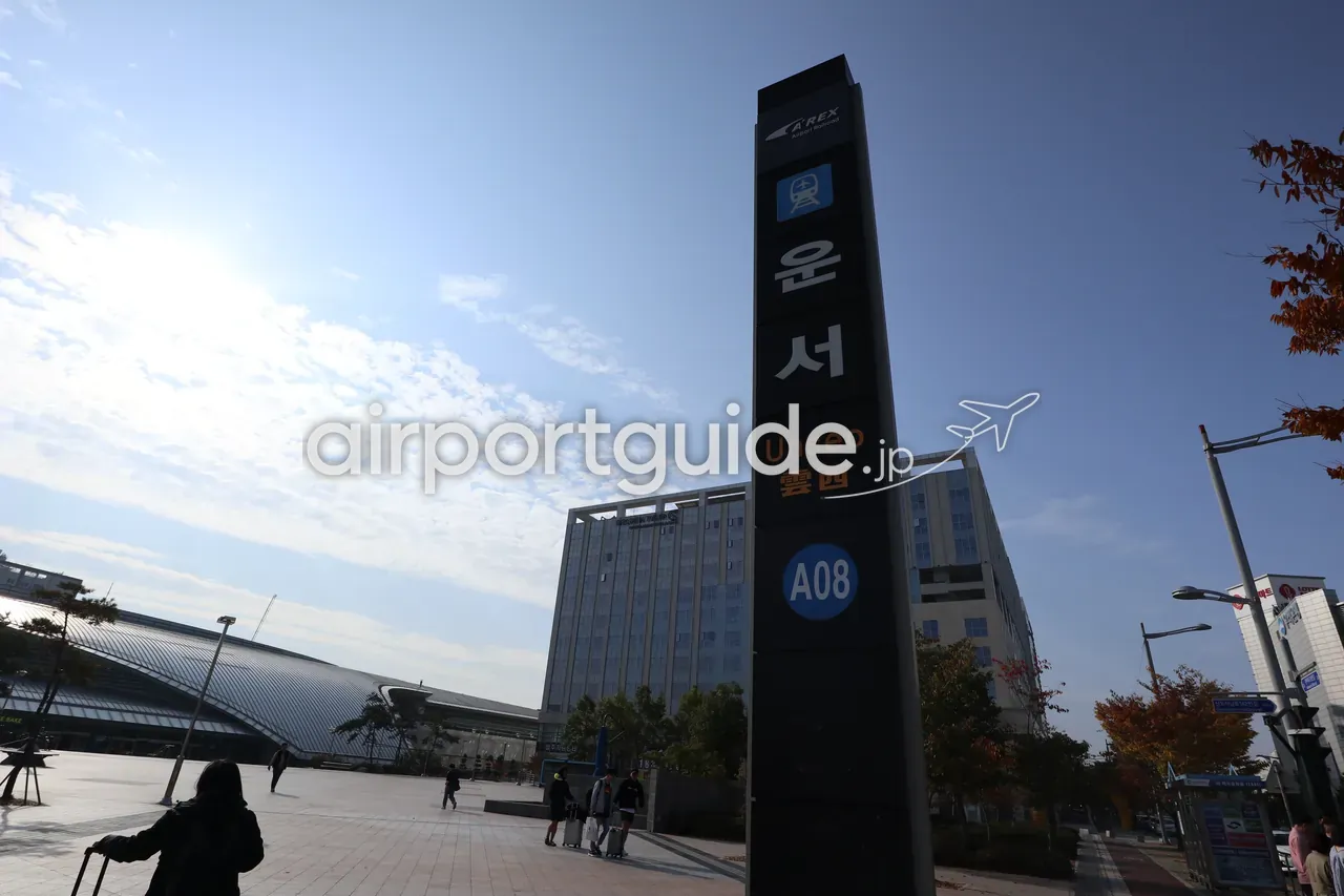空港鉄道A'REX 雲西（Unseo）駅の駅名標ポール。背後に駅舎と広場、スーツケースを引く旅行者が写る。