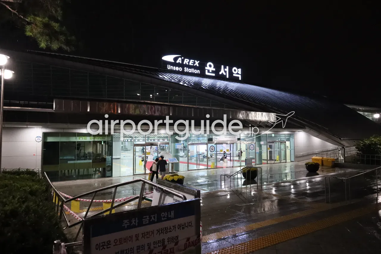 夜のA'REX 雲西（Unseo）駅の駅舎外観。曲面屋根の上に「A'REX Unseo Station 운서역」の発光サイン、ガラスの出入口、雨に濡れた床、傘の人。