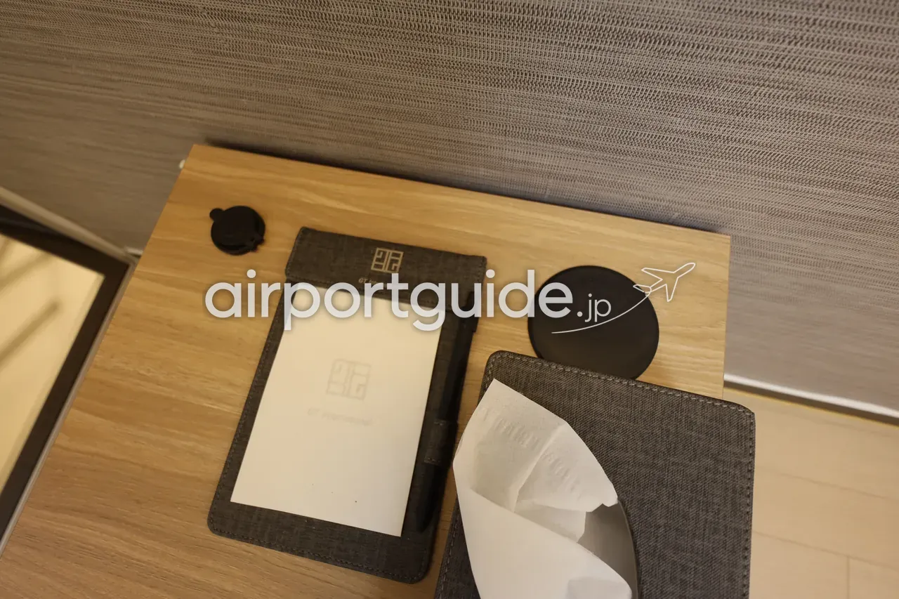 客室のサイドテーブルを俯瞰。「GT International」ロゴのグレーの案内フォルダーとペン、丸いワイヤレス充電パッド、布張りのティッシュボックス。壁は織り目の壁紙。