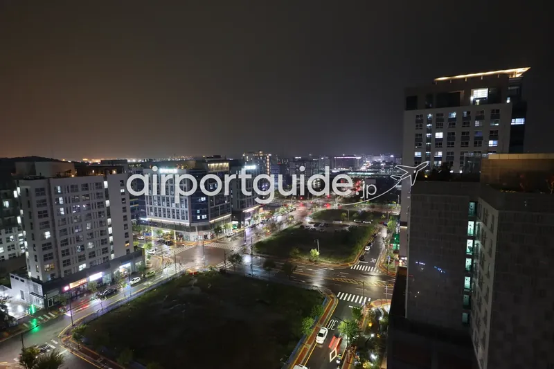 到着した夜の眺め。雨に濡れた大通りと交差点、街灯、遠くに空港方向の明かり。手前は造成途中の空き地。