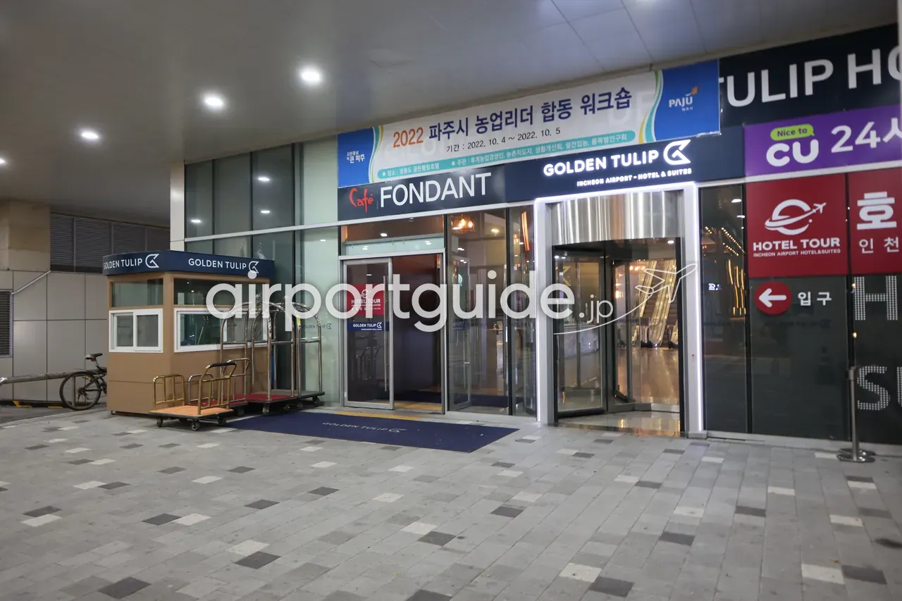 夜のゴールデンチューリップ仁川空港ホテルの玄関。「Café FONDANT」「GOLDEN TULIP INCHEON AIRPORT · HOTEL & SUITES」のサイン、回転扉、黄金色のベルカート2台、警備ボックス、右手にCU24時間コンビニと白いシャトルバン。